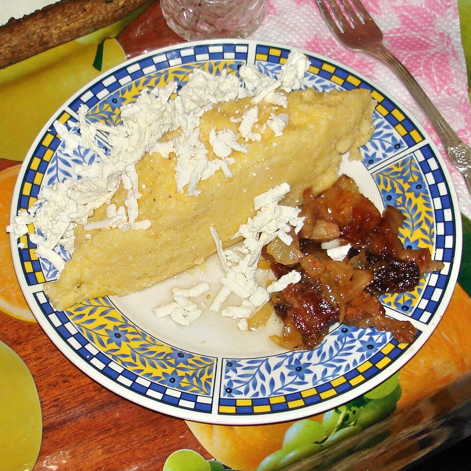 Una porción de mămăligă en un plato, cubierta con queso rallado, junto a cortezas de cerdo fritas